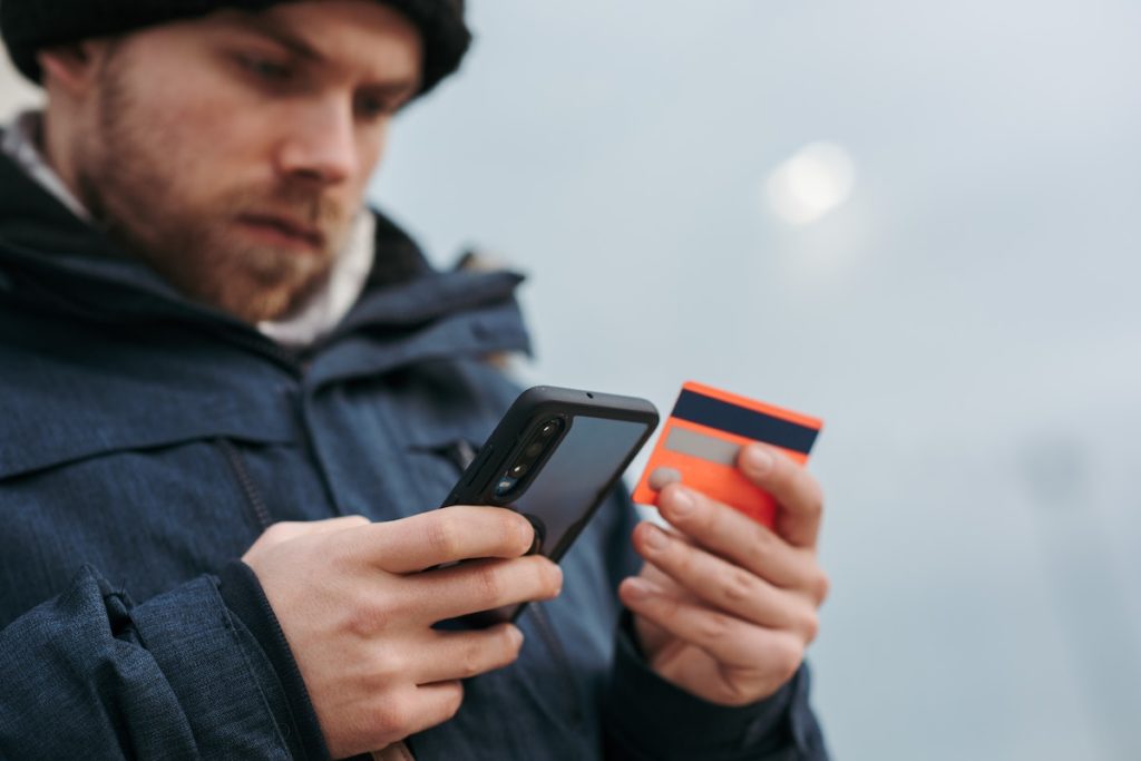 homem agasalhado segurando um celular com uma mão e um cartão de crédito com outra