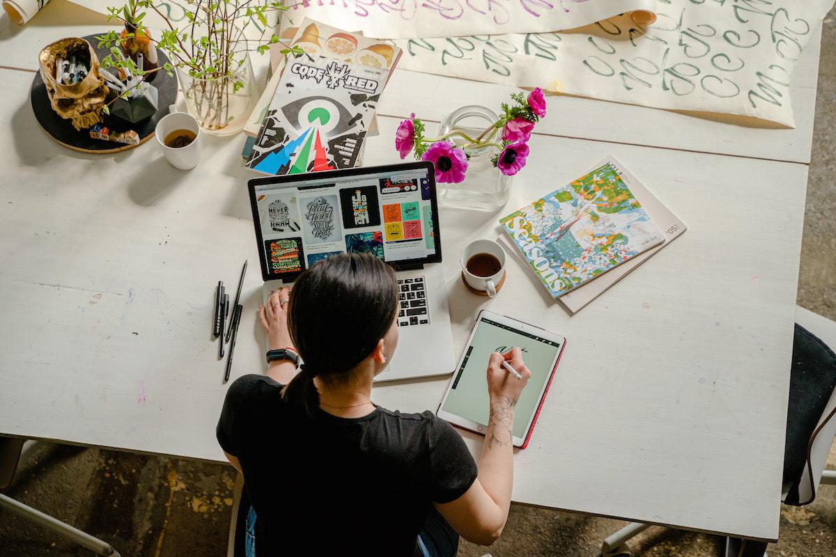 Homem trabalhando com notebook e tablet, que usa para desenhar, sobre mesa branca de madeira com objetos espalhados ao seu redor