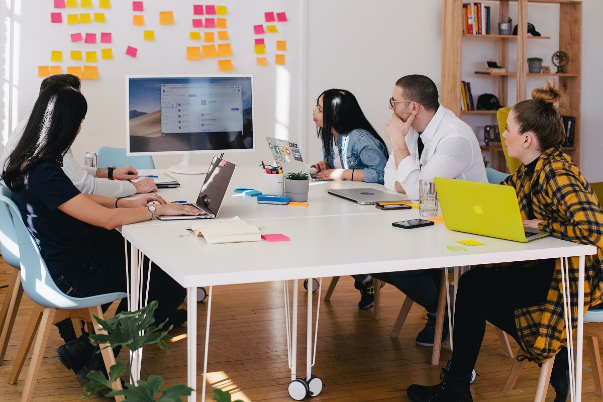 Pessoas em sala de reunião analisando dados em monitor diante de todos, que estão sentados ao redor de mesa, na frente de painel branco com vários post-its rosa e laranja colados no mesmo