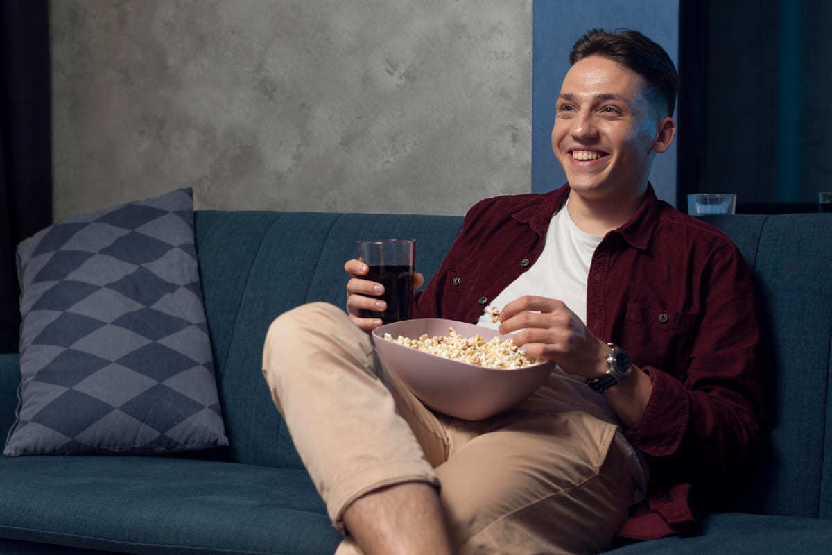 Homem sentado em sofá com bandeja de pipoca e copo de refrigerante de cola assistindo vídeo em Plataforma de streaming