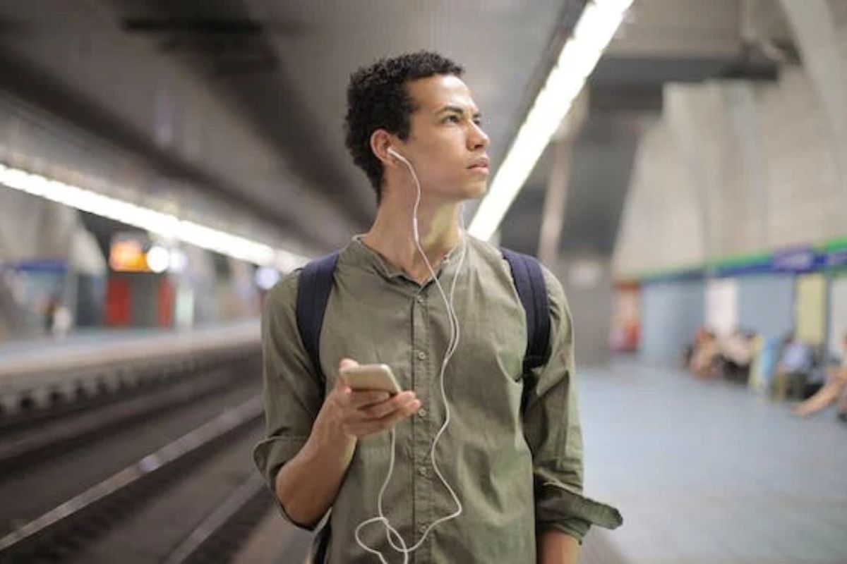 Homem em estação de metrô segurando celular com fone de ouvindo e ouvindo Podcasts