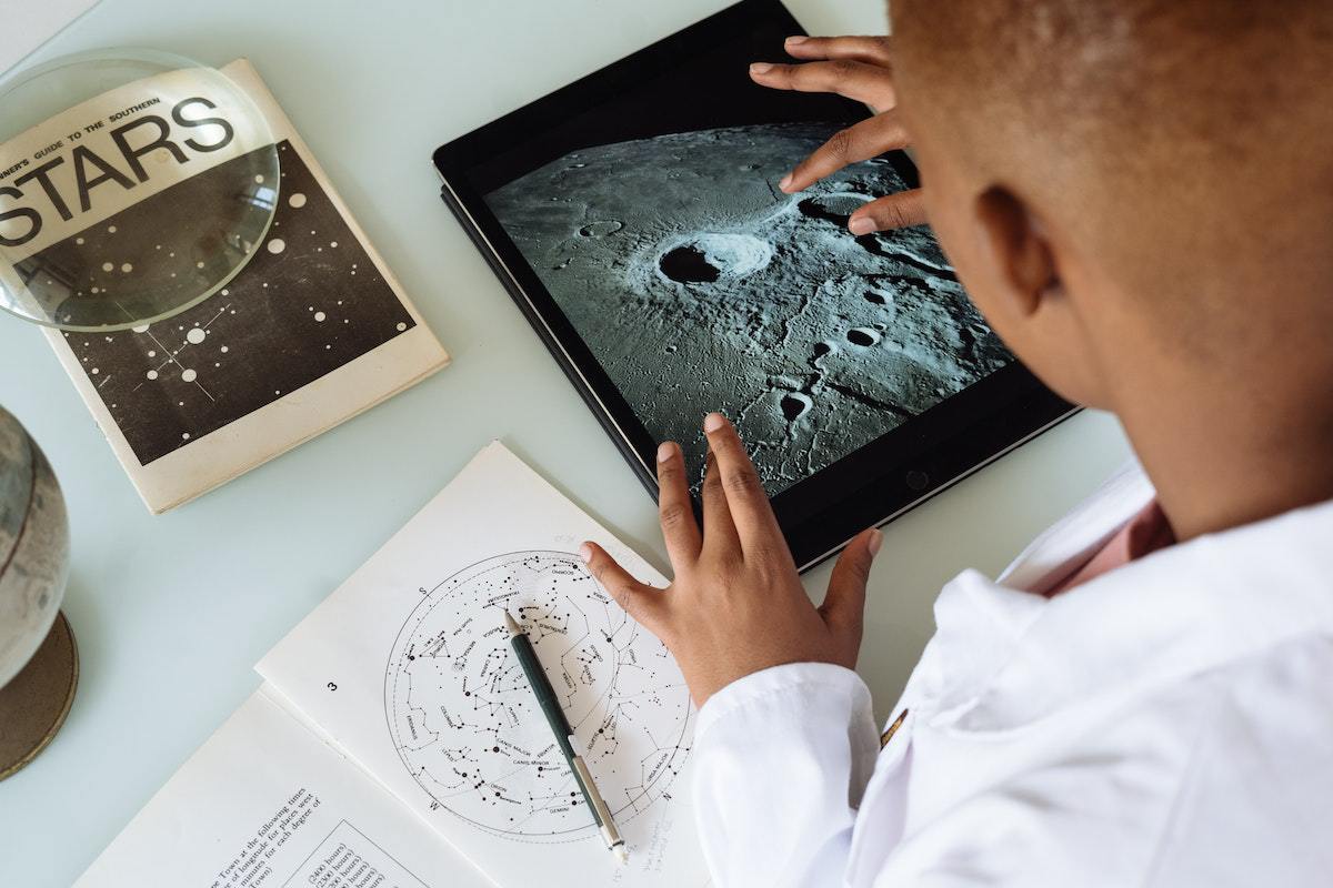 Homem analisando foto grande de terreno de local que parece ser a lua, com livros sobre astros ao seu redor, além de globo terrestre do tamanho médio de uma bola de futebol, com livro de cálculos à sua esquerda também, aparentemente de equações físicas