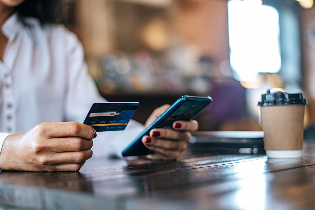 Pessoa usando Tecnologia bancária com celular na mão esquerda e cartão de banco à direita em ambiente interno de local que parece restaurante com copo de café ao seu lado sobre superfície de madeira