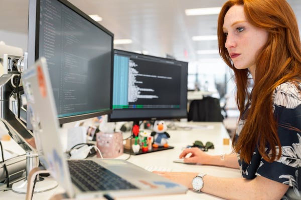 Mulher programando analisando código fonte em computador e notebook com dois monitores sobre mesa de madeira em local de trabalho