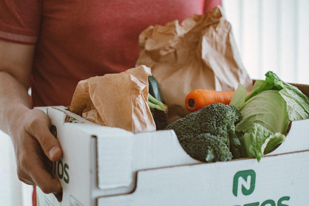 Pessoa se locomovendo enquanto carrega caixa com legumes e verduras
