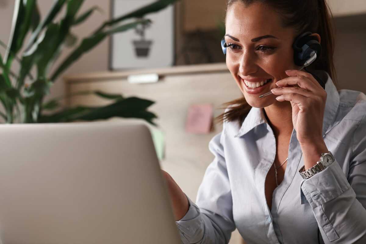 Mulher em local de trabalho sentada diante de notebook sorridente enquanto usa fone tipo headset, mas unilateral, com microfone para atendimentos