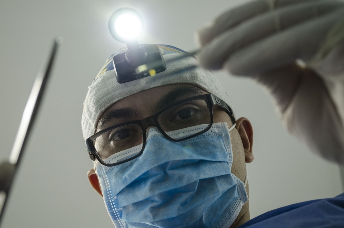 Homem, aparentemente médico, com capuz com lanterna forte e instrumentos cirúrgicos na mão, como se estivesse se debruçando para fazer procedimento em paciente