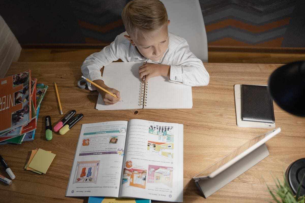 Menino fazendo suas atividades de casa escrevendo algo em caderno sobre mesa de madeira e usando livro e um tablet apoiado em suporte, uma das Tecnologias na educação infantil que mais ajudam no desenvolvimento, com outros livros e lápis e canetas, bem como agenda, ao seu redor em cima dessa mesa