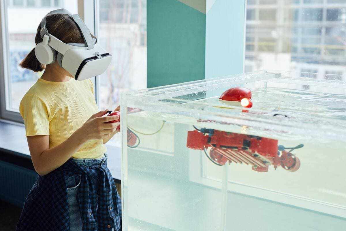 Criança de pé diante de reservatório grande e transparente de água usando óculos de realidade virtual como uma das Tecnologias na educação infantil e mexendo em objeto que lembra joystick