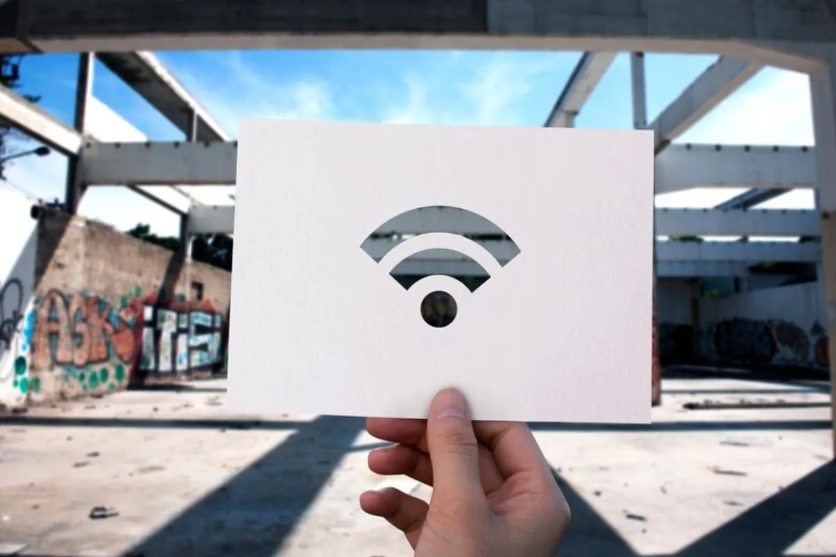 Símbolo do wi-fi representado em imagem de pessoa segurando papel com esse grafismo em meio à cenário urbano semelhante à praça.