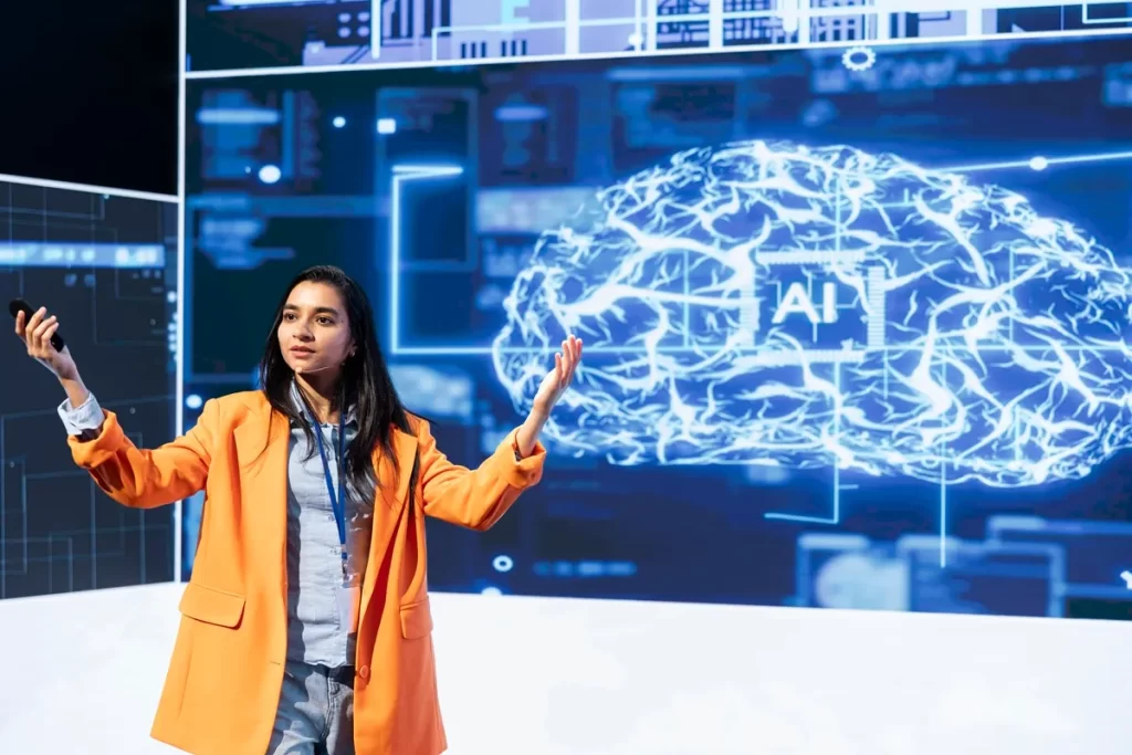 Mulher apresentando conceitos de Inteligência Artificial em um palco, com uma imagem digital de um cérebro iluminado e a palavra "AI" projetada ao fundo.