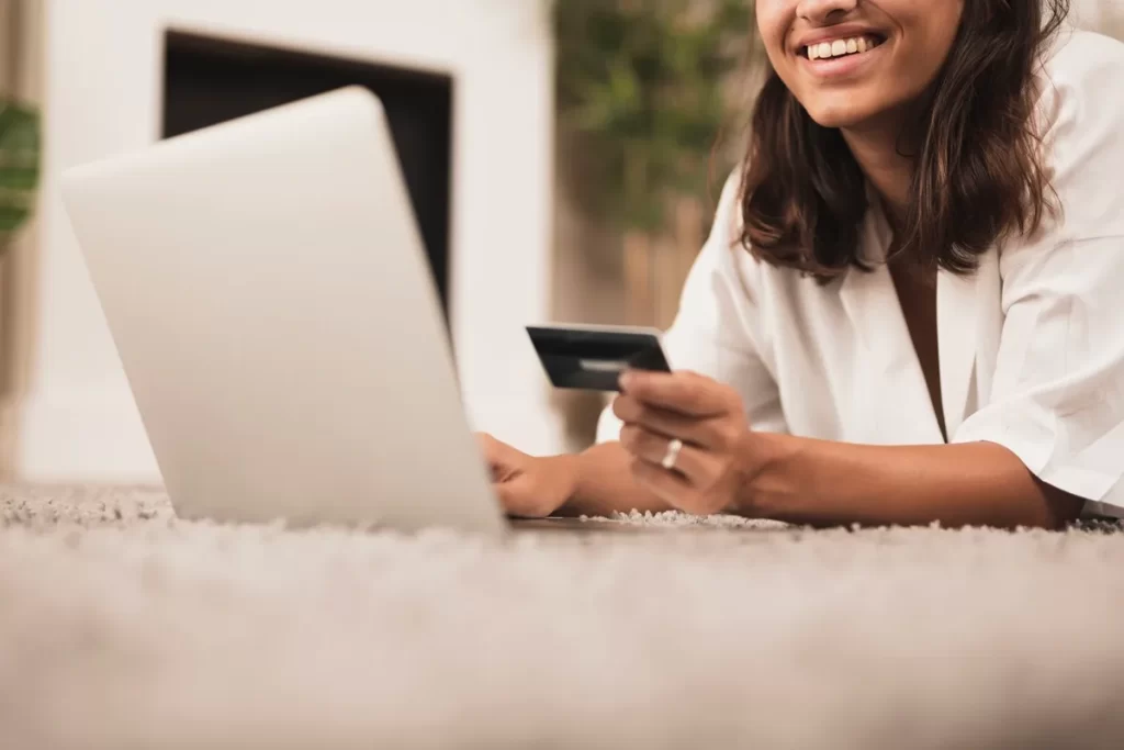 Mulher sorrindo enquanto realiza compras online com um cartão de crédito em mãos e usando um laptop sobre um tapete felpudo.