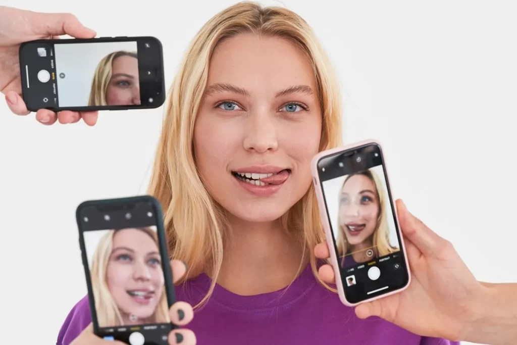 Mulher jovem de cabelos loiros sorrindo, enquanto várias mãos seguram smartphones capturando selfies dela em ângulos diferentes.