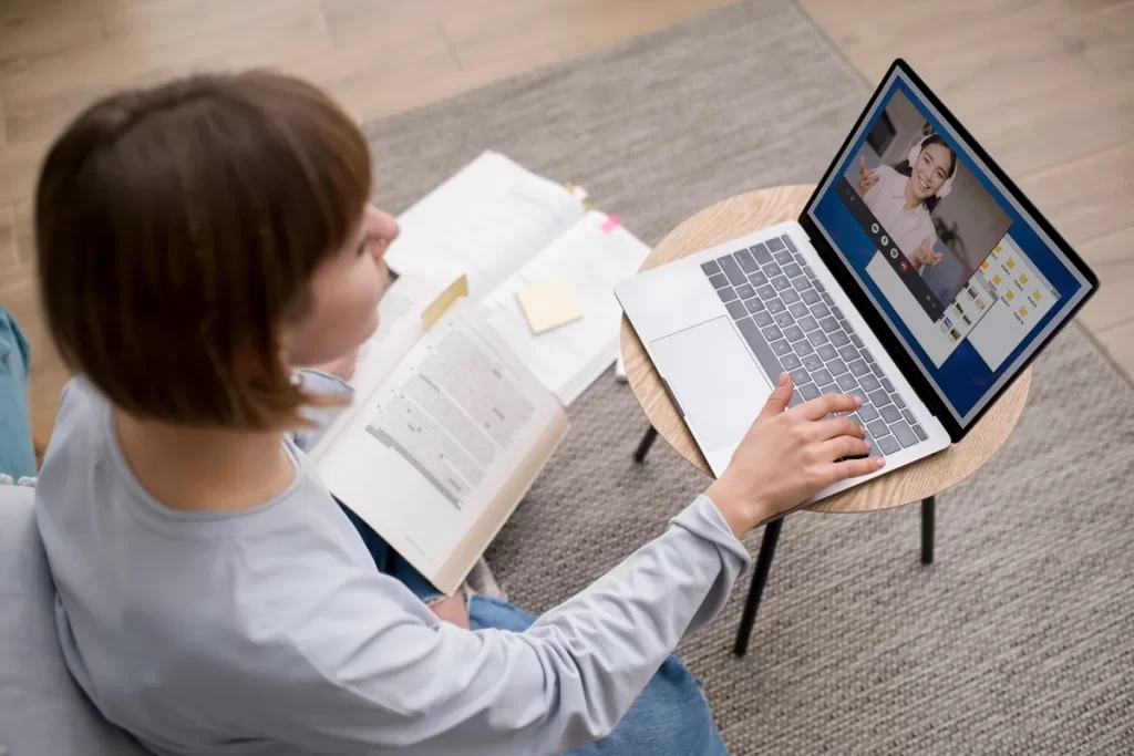 Pessoa participando de uma videoconferência em um laptop enquanto segura um livro aberto, em um ambiente de estudo acolhedor, com materiais organizados sobre uma mesa.