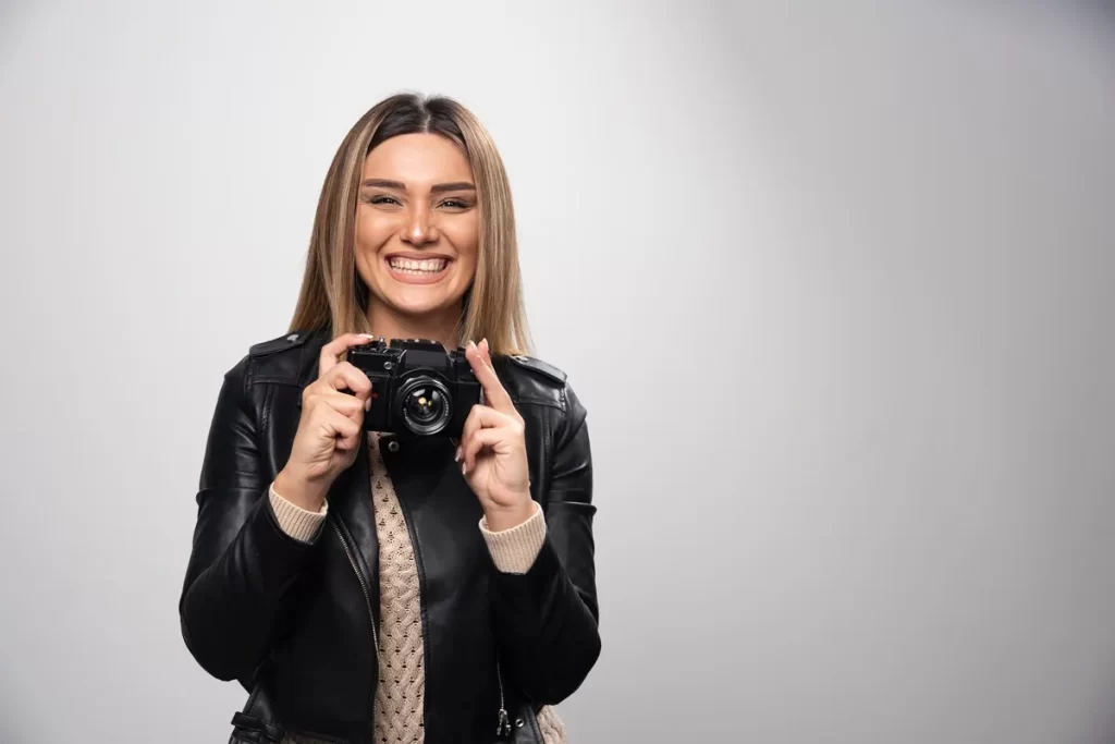 Mulher sorridente segurando uma câmera fotográfica em um fundo cinza neutro, vestindo jaqueta de couro preta e suéter claro.