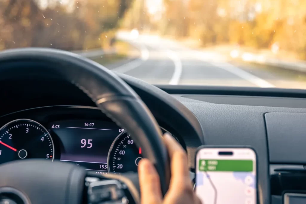 Imagem de um motorista dirigindo com uma mão no volante enquanto utiliza um GPS no smartphone durante uma viagem em estrada aberta.