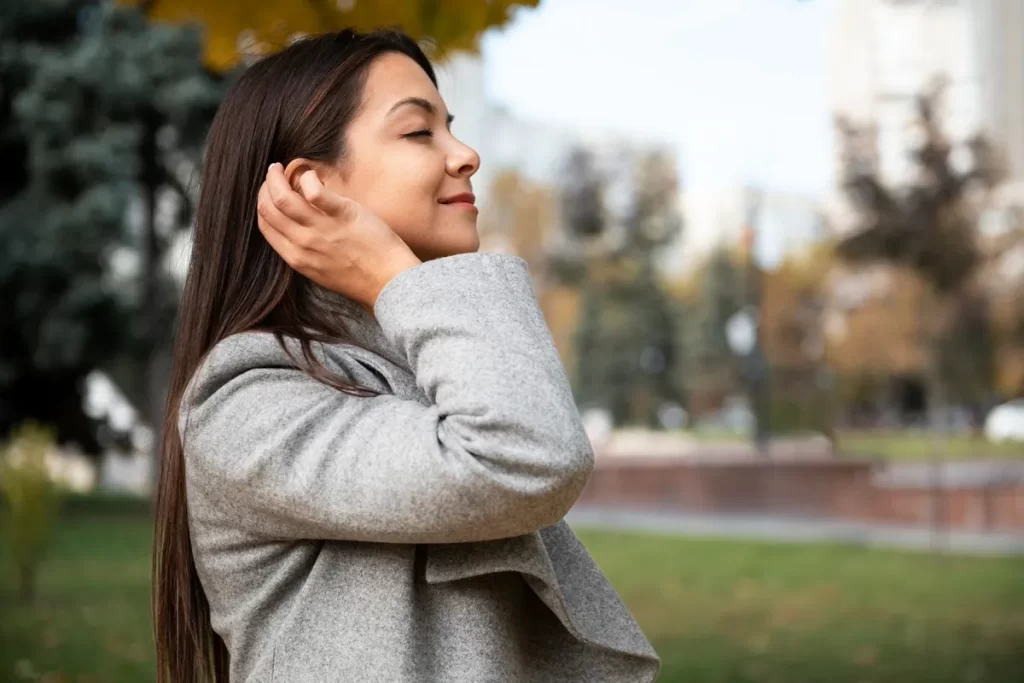 Mulher com expressão serena, vestindo casaco cinza, aproveitando o ar livre em um parque com árvores e grama ao fundo, representando relaxamento e conexão com a natureza.