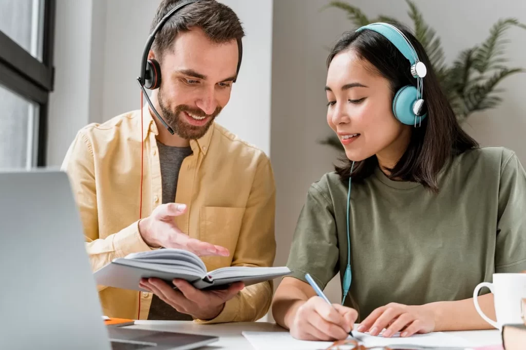 Dupla de profissionais com fones de ouvido colaborando em um ambiente de trabalho. Um homem de camisa amarela explica algo mostrando um livro, enquanto uma mulher de fones azuis anota informações em uma folha de papel.