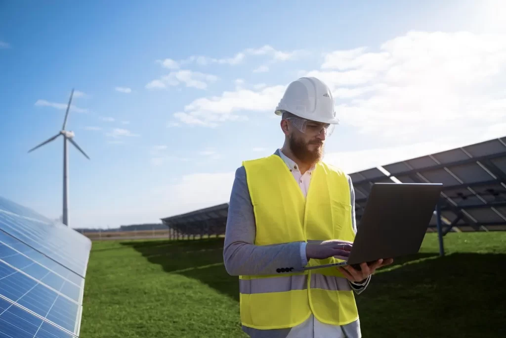 Engenheiro com colete de segurança amarelo e capacete branco utilizando um laptop em uma usina de energia solar, com turbinas eólicas ao fundo.