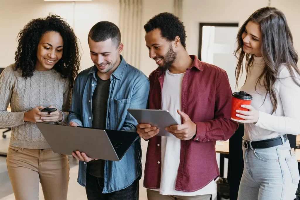 Grupo de jovens em um ambiente de trabalho colaborativo compartilhando informações em dispositivos eletrônicos, como notebook e tablet.