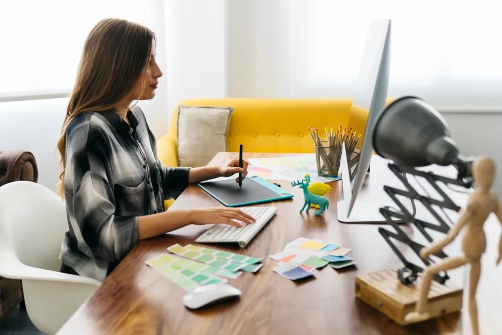 Designer gráfico trabalhando em um computador com mesa digitalizadora, paletas de cores e lápis de desenho, em um ambiente criativo e iluminado.